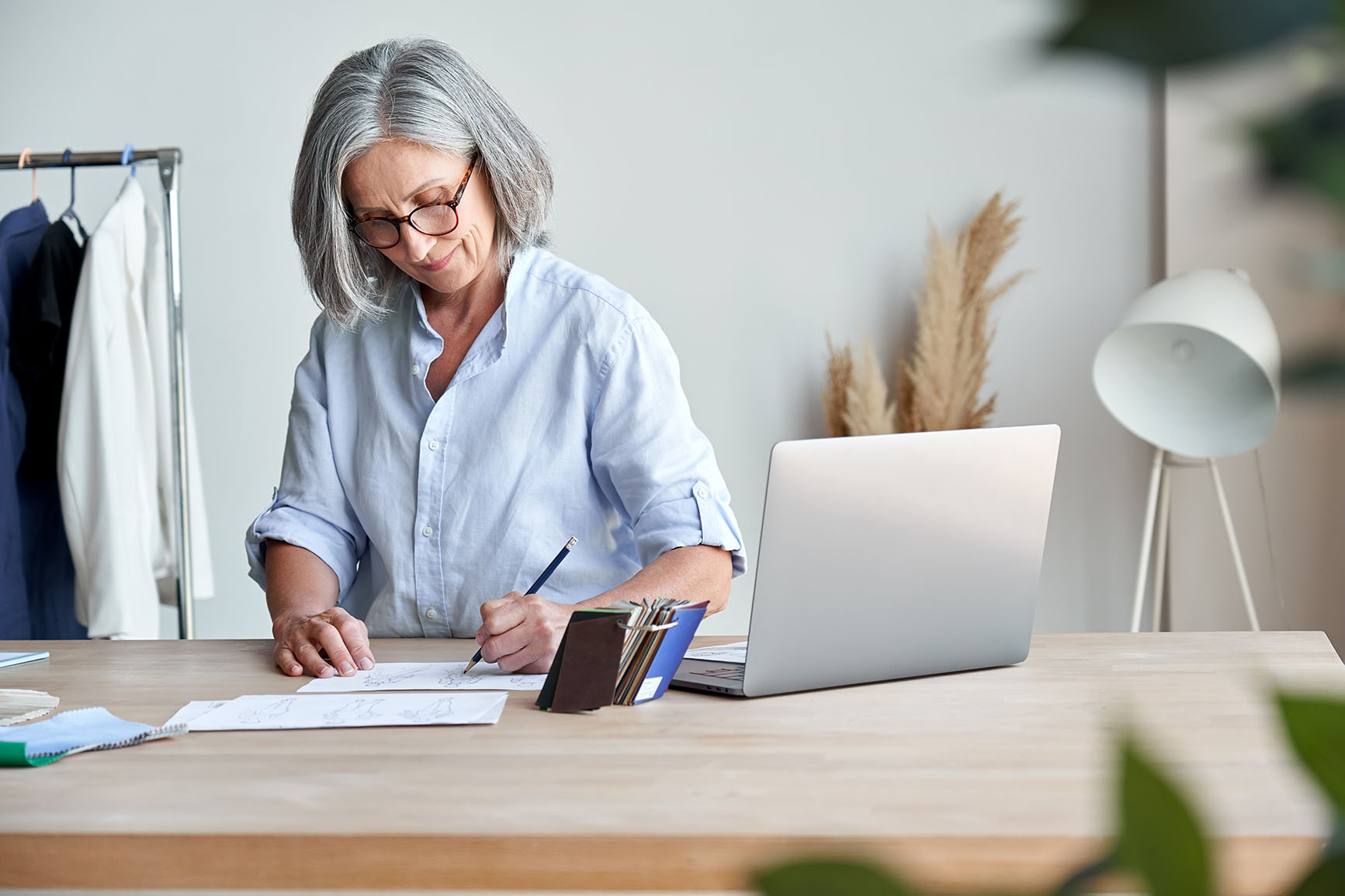 An older woman sketching at a desk with her laptop next to her